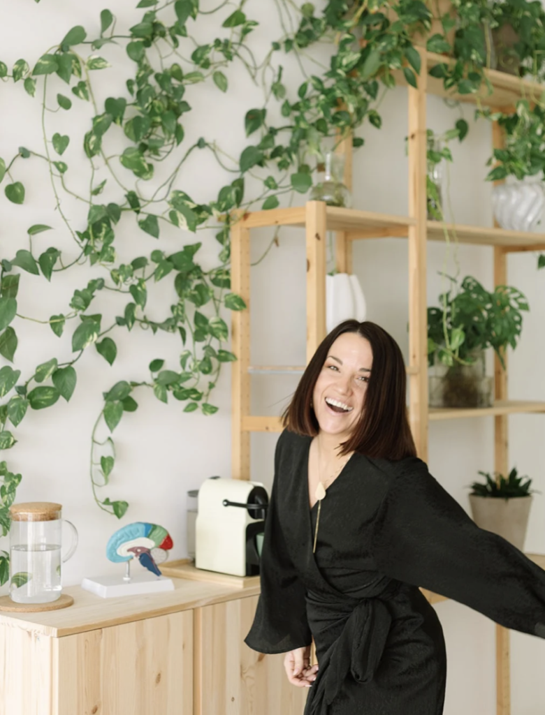 Gladys Kali sonriendo en su estudio de neurociencia, junto a plantas, una cafetera y un modelo anatómico del cerebro.